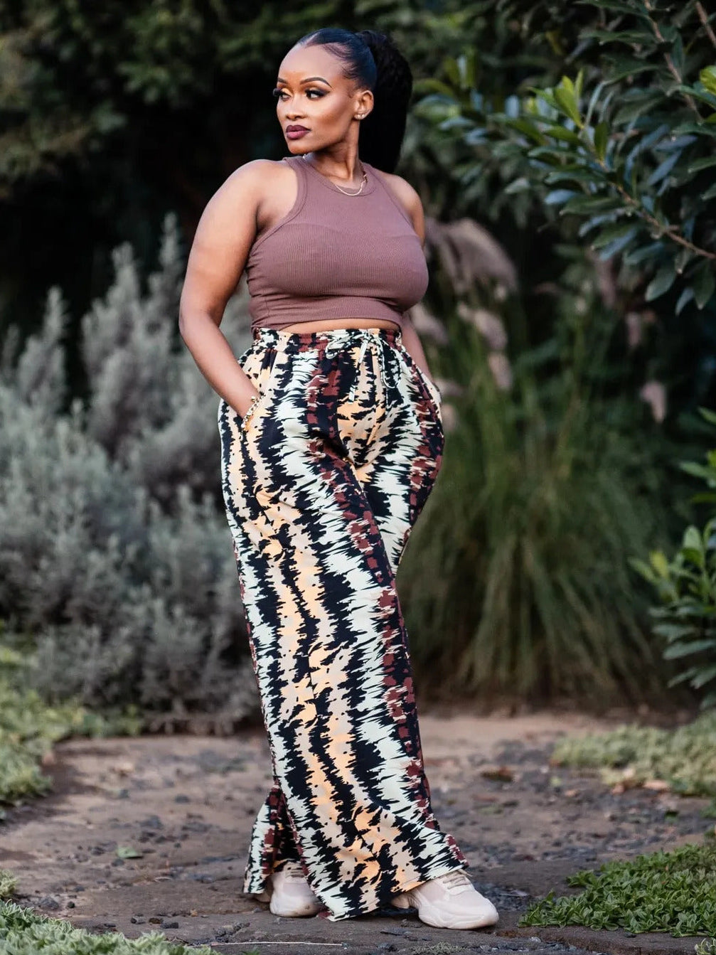 Woman wearing a brown top and patterned pants standing outdoors with greenery in the background