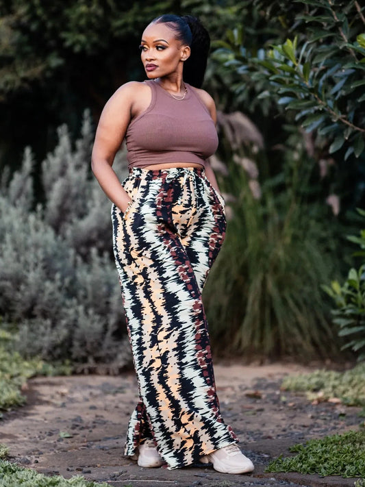 Woman wearing a brown top and patterned pants standing outdoors with greenery in the background